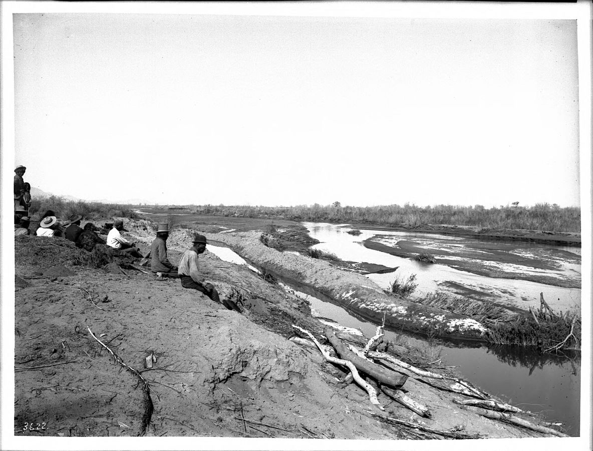 Pima Indian farmers sitting on shore looking at an irrigation dam on the Gila River, Arizona around 1900